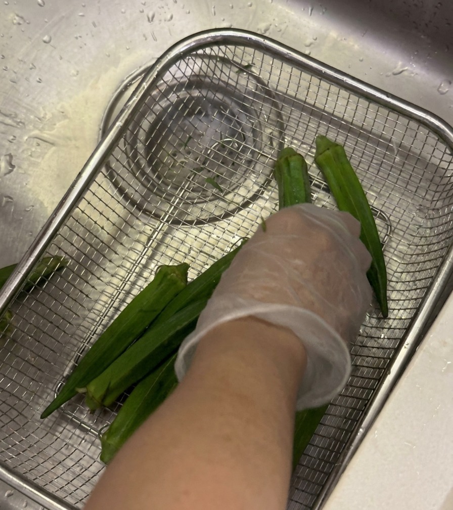 Vainas de okra fresca siendo lavadas en una cesta de malla bajo el grifo mientras una mano enguantada frota las piezas.