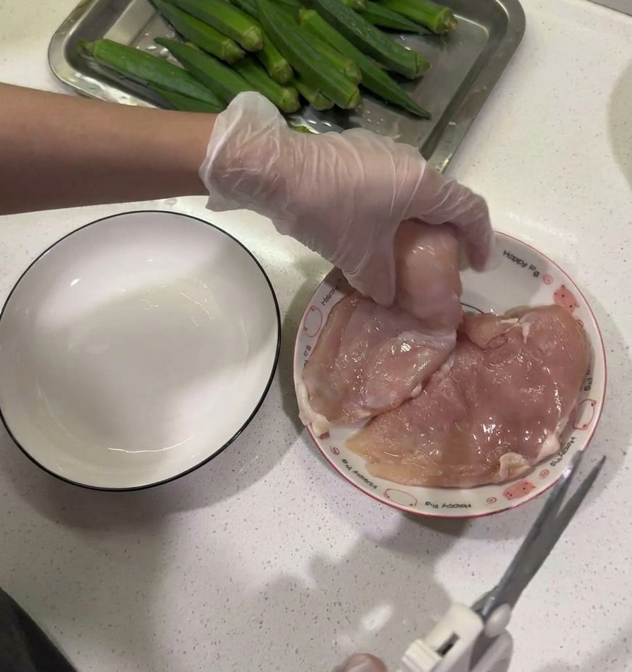 Tijeras de cocina recortando la grasa de una pechuga de pollo cruda sobre un plato junto a un cuenco vacío.