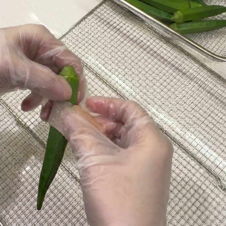 Manos con guantes enrollando una lámina fina de pollo crudo en espiral alrededor de una vaina de okra verde entera.