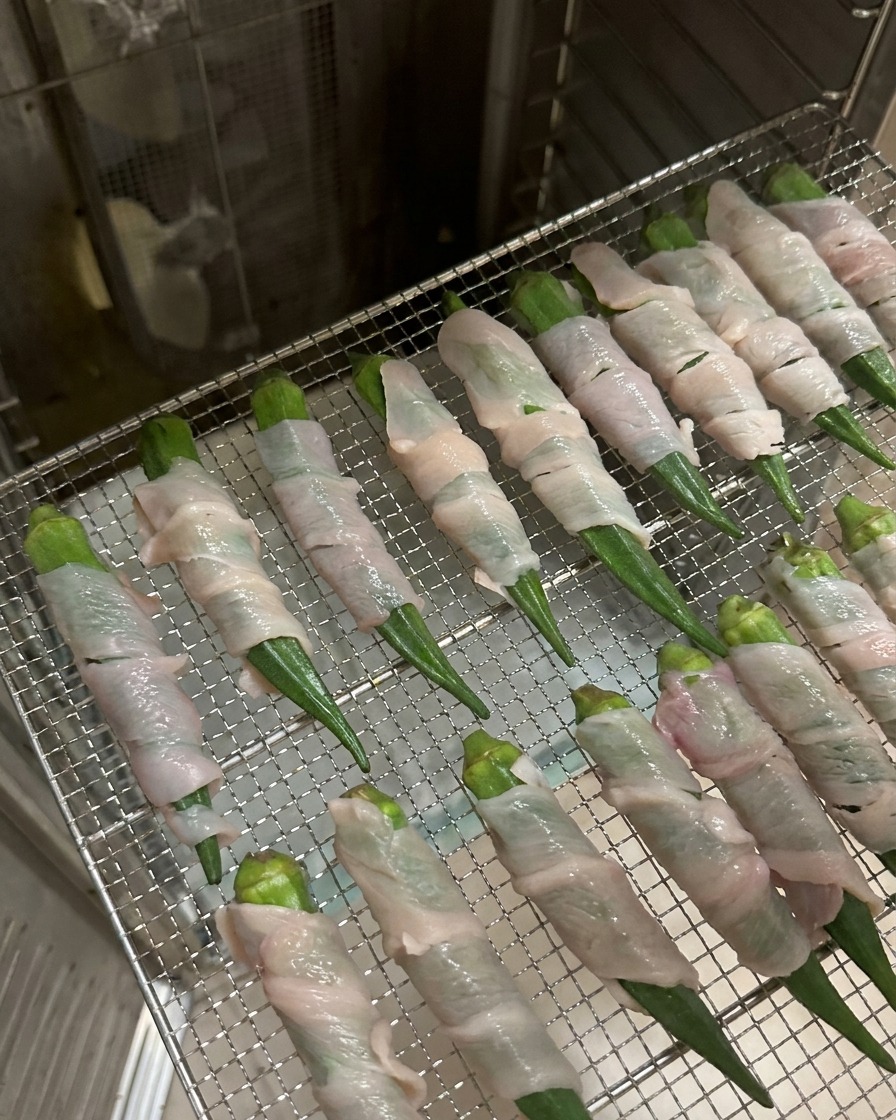 Filas de okra envuelta en pollo dispuestas sobre una rejilla metálica dentro de un deshidratador de acero inoxidable.