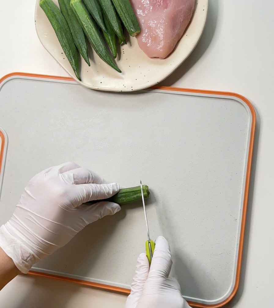 Manos con guantes usando un cuchillo pequeño para cortar los extremos de vainas de okra verde sobre una tabla de cortar blanca.