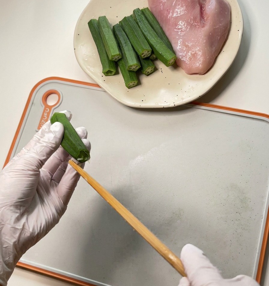 Una persona con guantes blancos utiliza un palillo de madera para extraer las semillas y la pulpa de una vaina de okra verde.