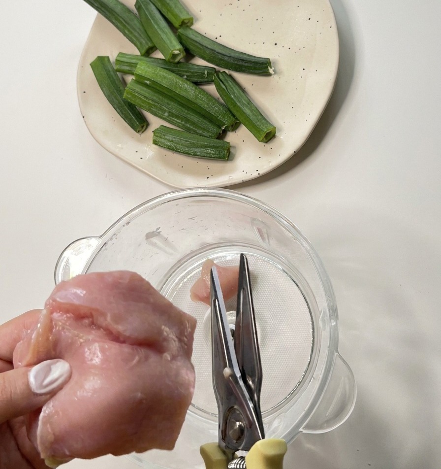 Manos usando tijeras de cocina de metal para cortar pechuga de pollo cruda dentro de una licuadora transparente.
