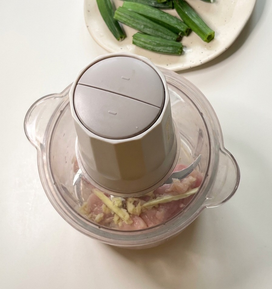 Un procesador de alimentos sobre una encimera de cocina con trozos de pollo crudo y semillas de okra, listo para procesar.