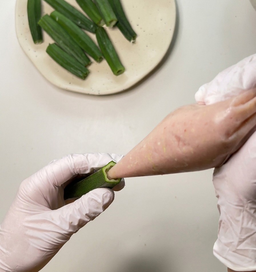 Un primer plano de una persona introduciendo pasta de pollo rosada en una vaina de okra verde y hueca usando una manga pastelera.