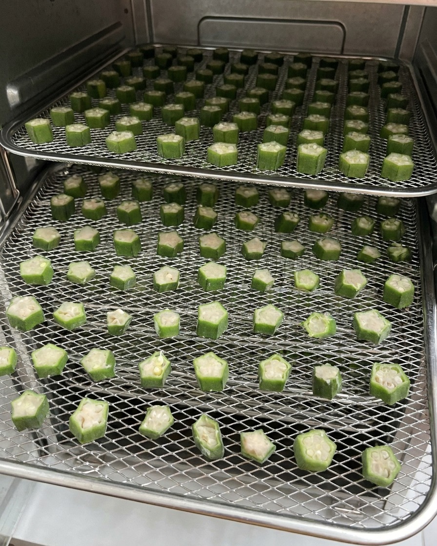 Dos rejillas de alambre llenas de premios de okra y pollo cortados en rodajas, colocadas dentro de un deshidratador de alimentos durante el proceso de secado.