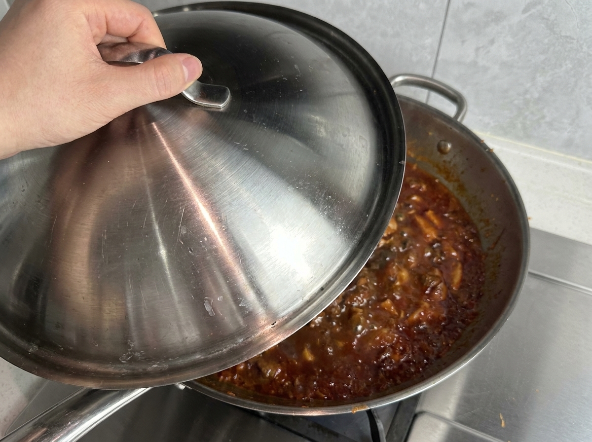 Mano colocando una tapa de acero inoxidable sobre una sartén tipo wok llena de salsa roja burbujeante.