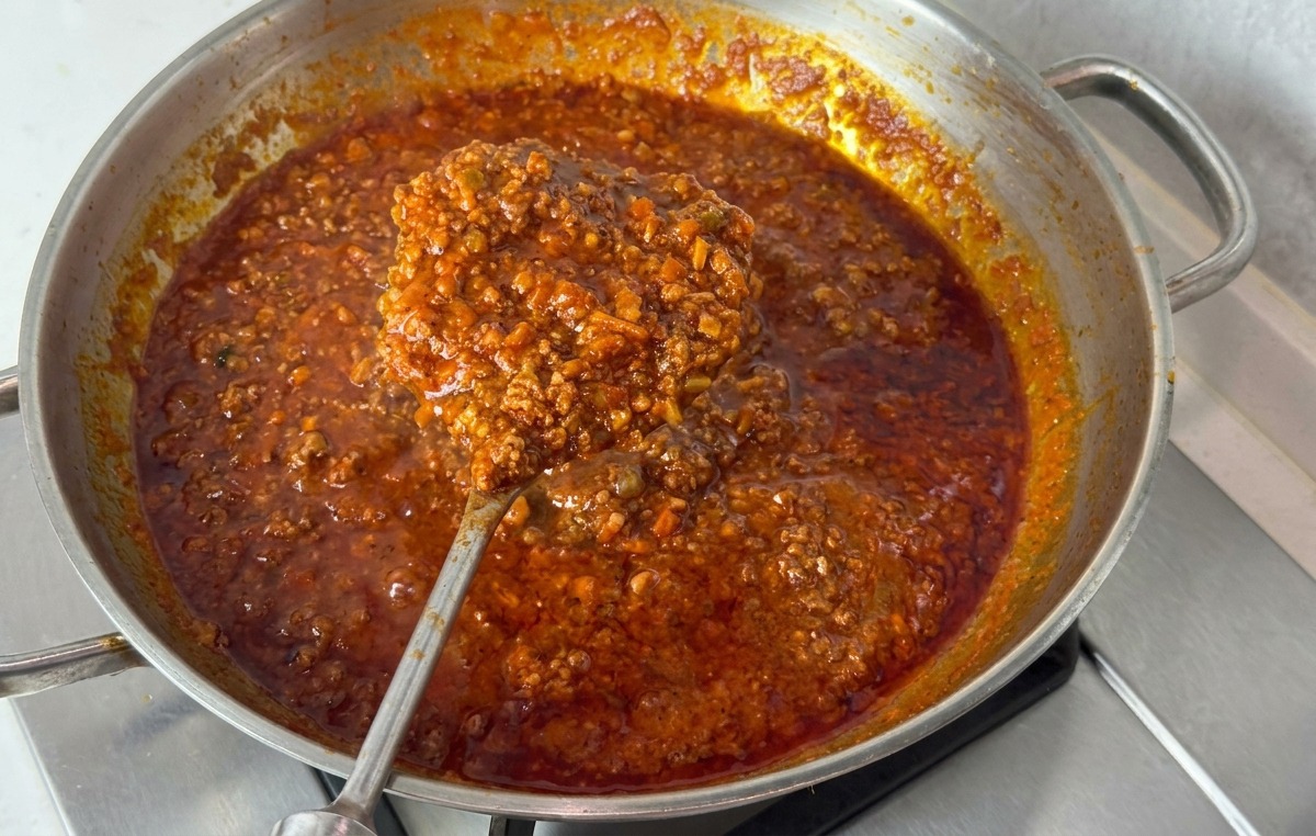 Sartén de acero con salsa boloñesa oscura y espesa siendo levantada con una cuchara para mostrar su consistencia viscosa.