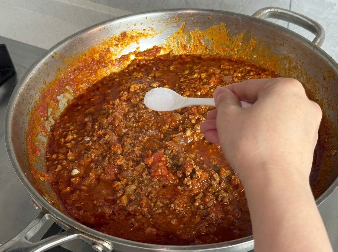 Mano con una cuchara pequeña de plástico llena de sal fina sobre la salsa de carne roja hirviendo.