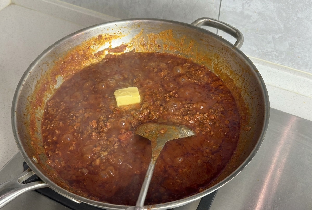 Bloque cuadrado de mantequilla amarilla reposando sobre la superficie de la salsa boloñesa en una sartén grande.
