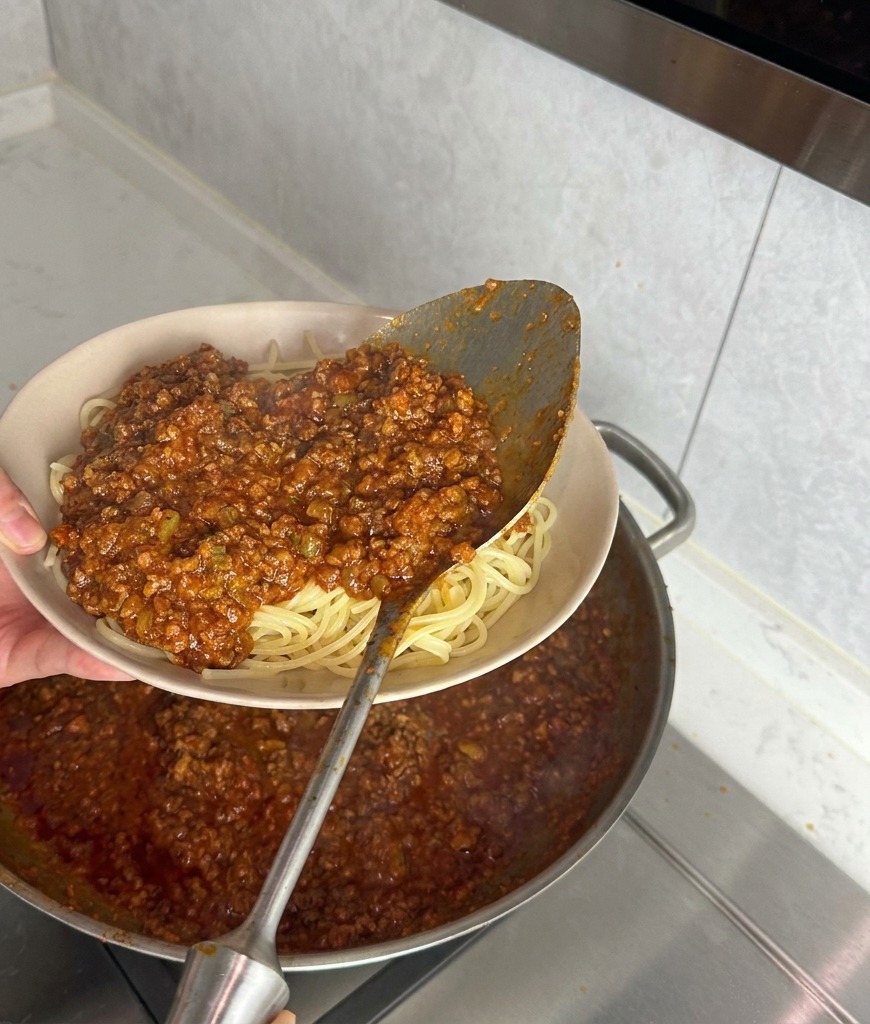 Cucharón de metal vertiendo salsa de carne espesa sobre una cama de espaguetis blancos en un bol beige.