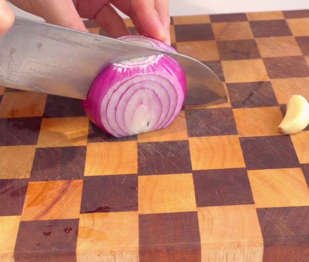 Una persona picando finamente una cebolla roja en cubos pequeños y uniformes sobre una tabla de madera.