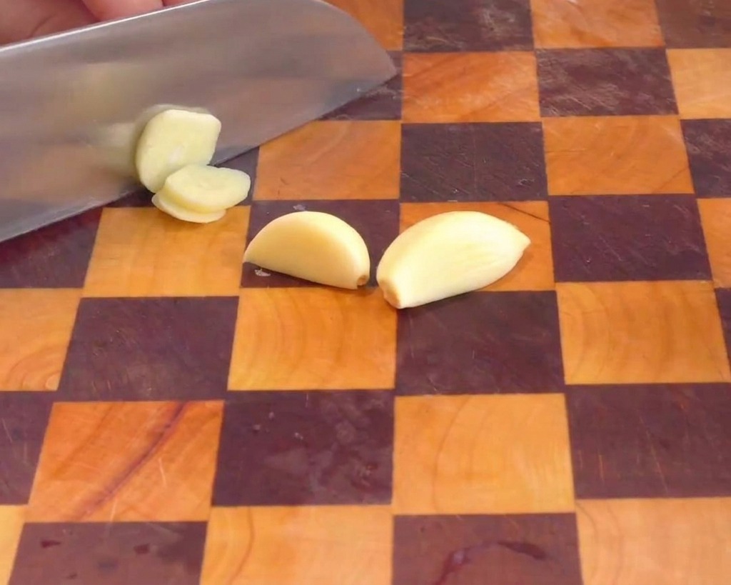 Una mano usando un cuchillo de chef para cortar dientes de ajo pelados en láminas finas sobre una tabla de madera.