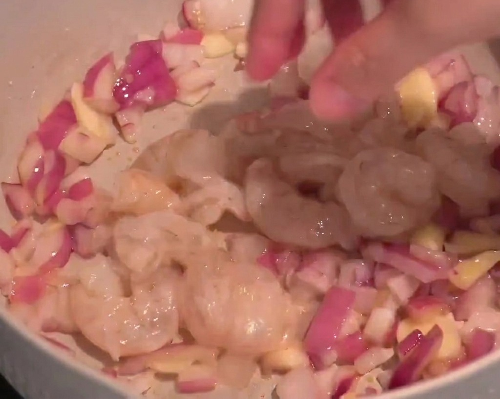 Camarones crudos translúcidos siendo añadidos a una sartén con cebolla roja y láminas de ajo.