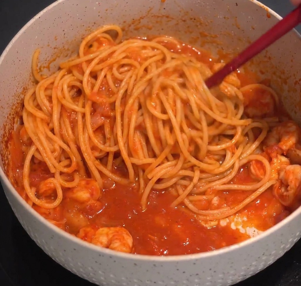 Espaguetis mezclándose con salsa roja de camarones usando pinzas de cocina en una olla moteada.