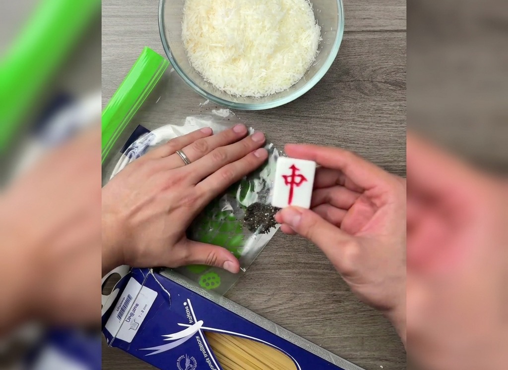Una mano usando el lado plano de una ficha de mahjong para triturar granos de pimienta dentro de una bolsa de plástico sobre una mesa de madera.