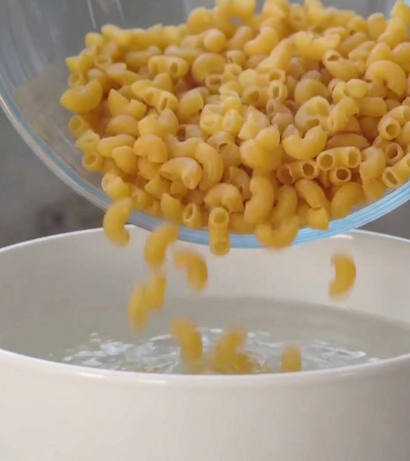 Espirales de pasta secos cayendo desde un bol de cristal a una olla blanca con agua hirviendo