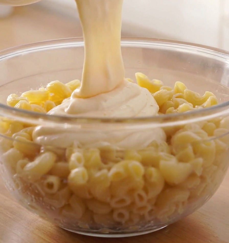 Un chorro espeso de salsa de queso fundido cayendo sobre un bol grande de pasta de espirales cocida