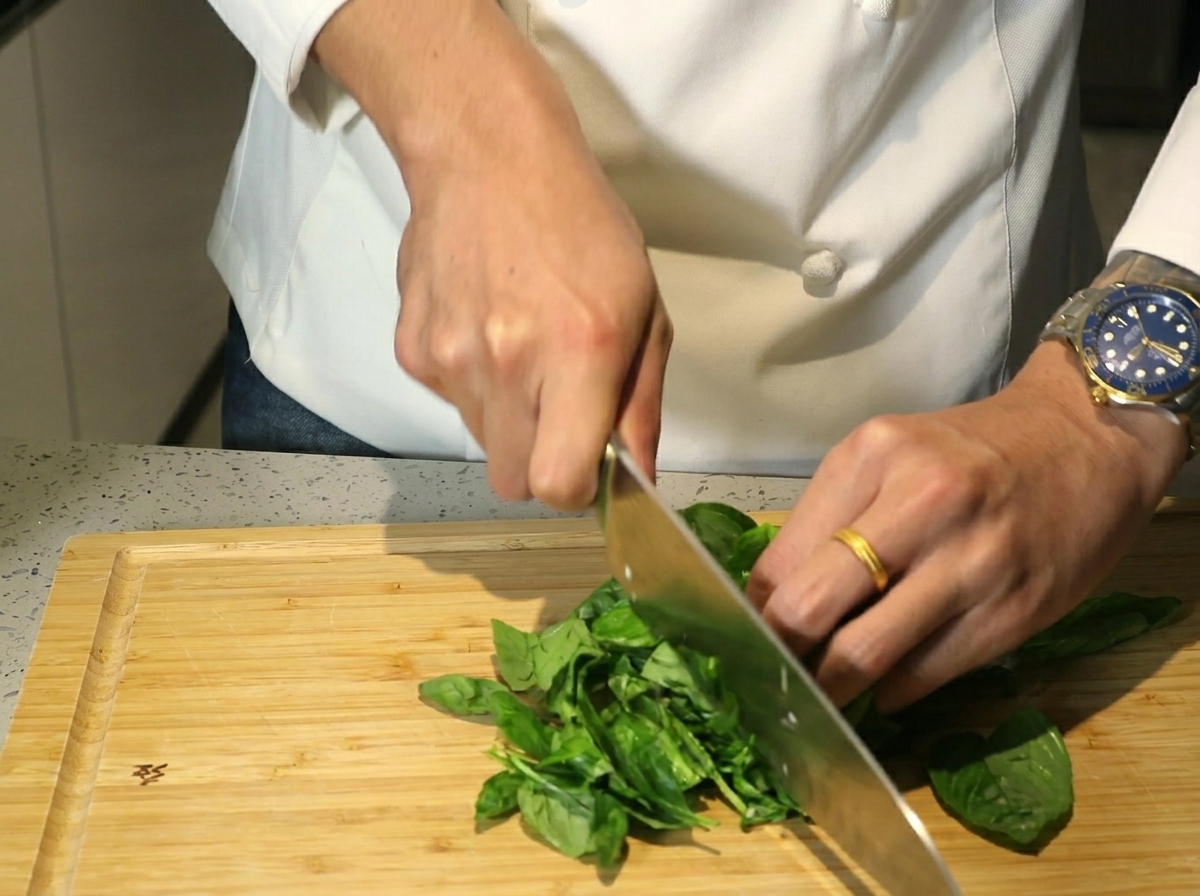 Manos usando un cuchillo grande para picar hojas de albahaca fresca sobre una tabla de madera.
