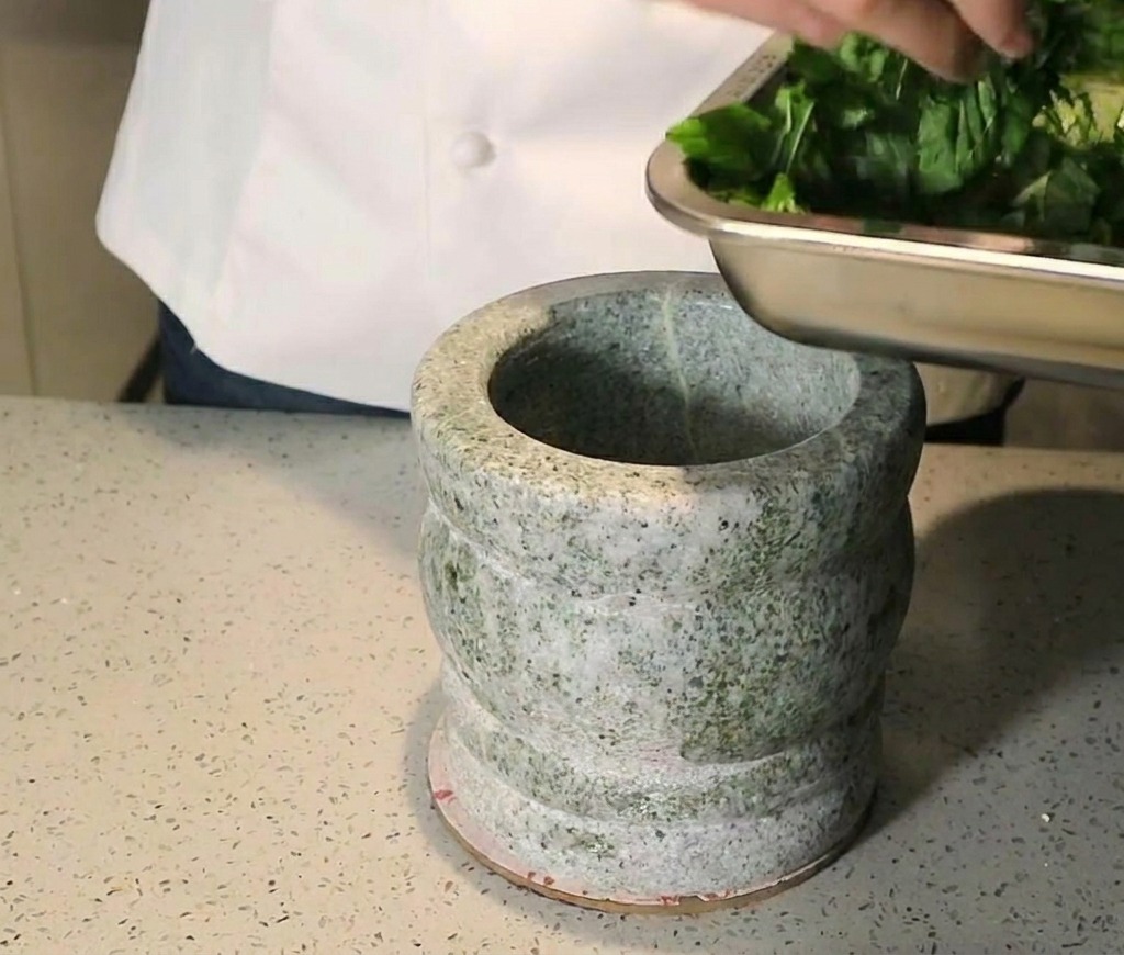 Una persona añadiendo hojas de albahaca picadas desde una bandeja metálica a un mortero de granito.