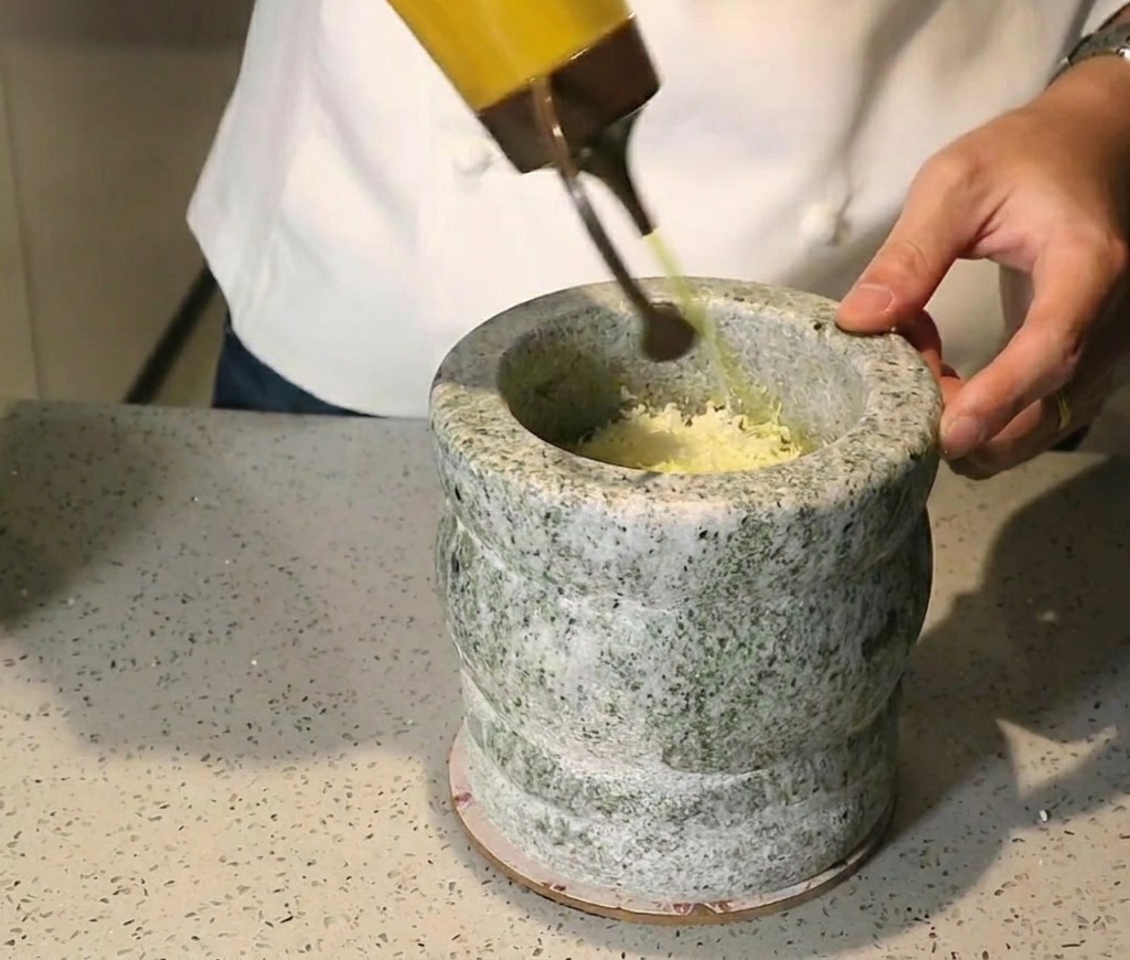 Aceite de oliva dorado vertiéndose desde un biberón de cocina en un mortero con hierbas y frutos secos.