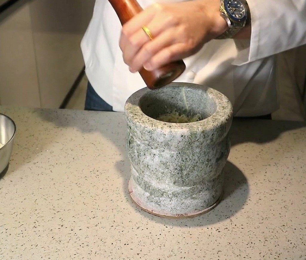 Un molinillo de madera moliendo pimienta negra sobre los ingredientes dentro de un mortero de piedra.