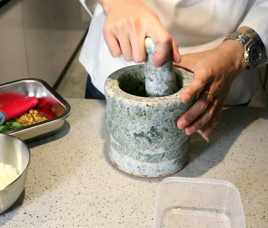 Un chef usando un mortero y maza de piedra para triturar albahaca y ajo hasta formar una pasta espesa.