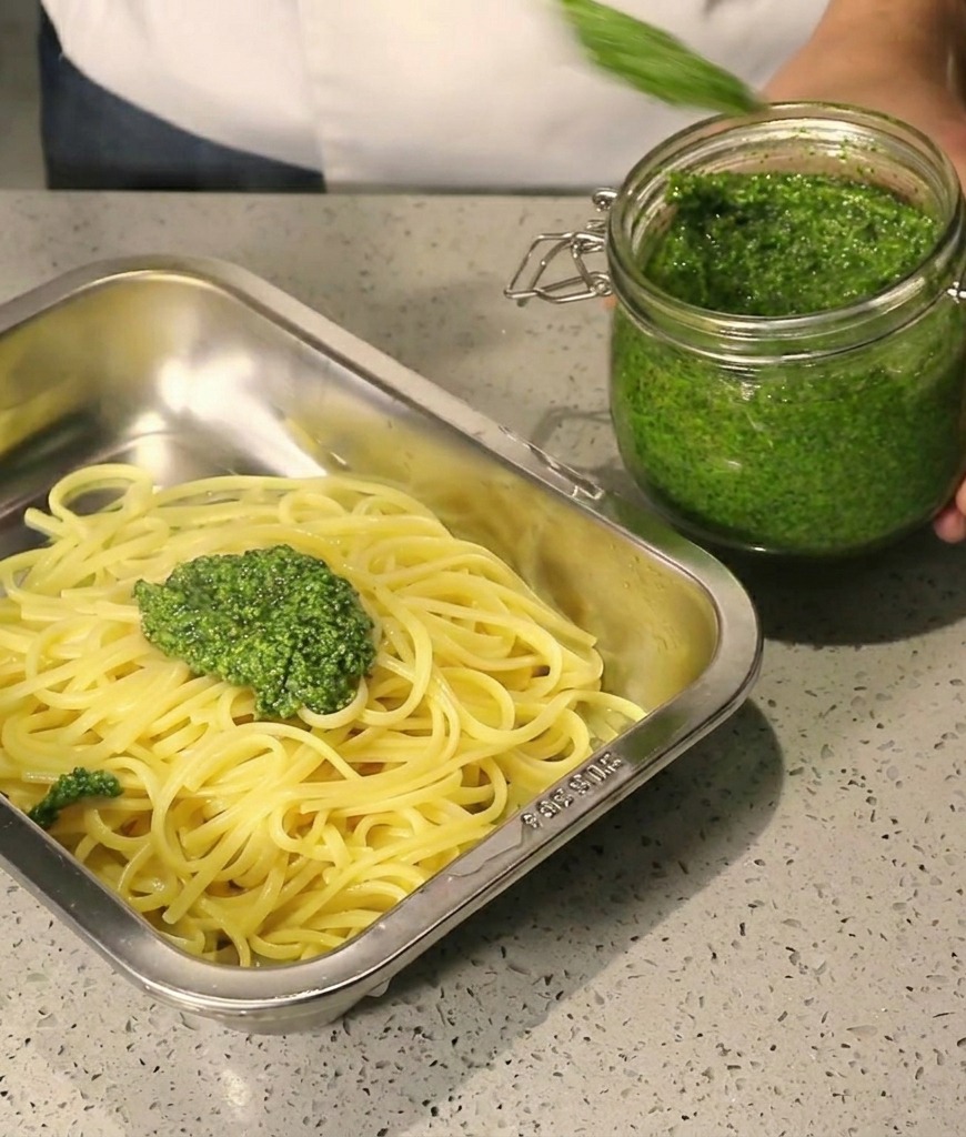 Salsa pesto verde espesa vertiéndose con una cuchara sobre espaguetis calientes en una bandeja de acero.