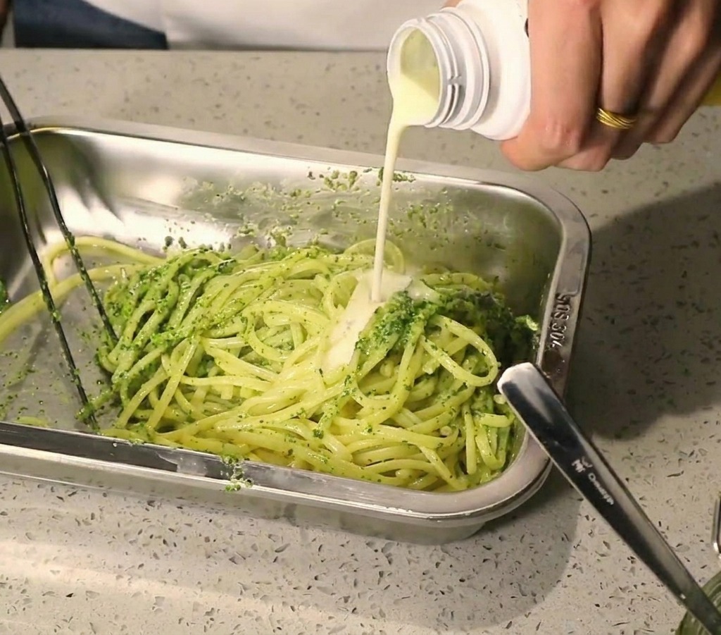 Un chorro de salsa blanca cremosa vertiéndose sobre la pasta mezclada con pesto verde.