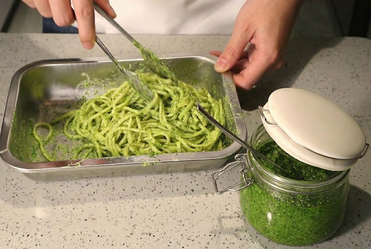 Usando pinzas metálicas para remover espaguetis con pesto y crema en una bandeja de metal.