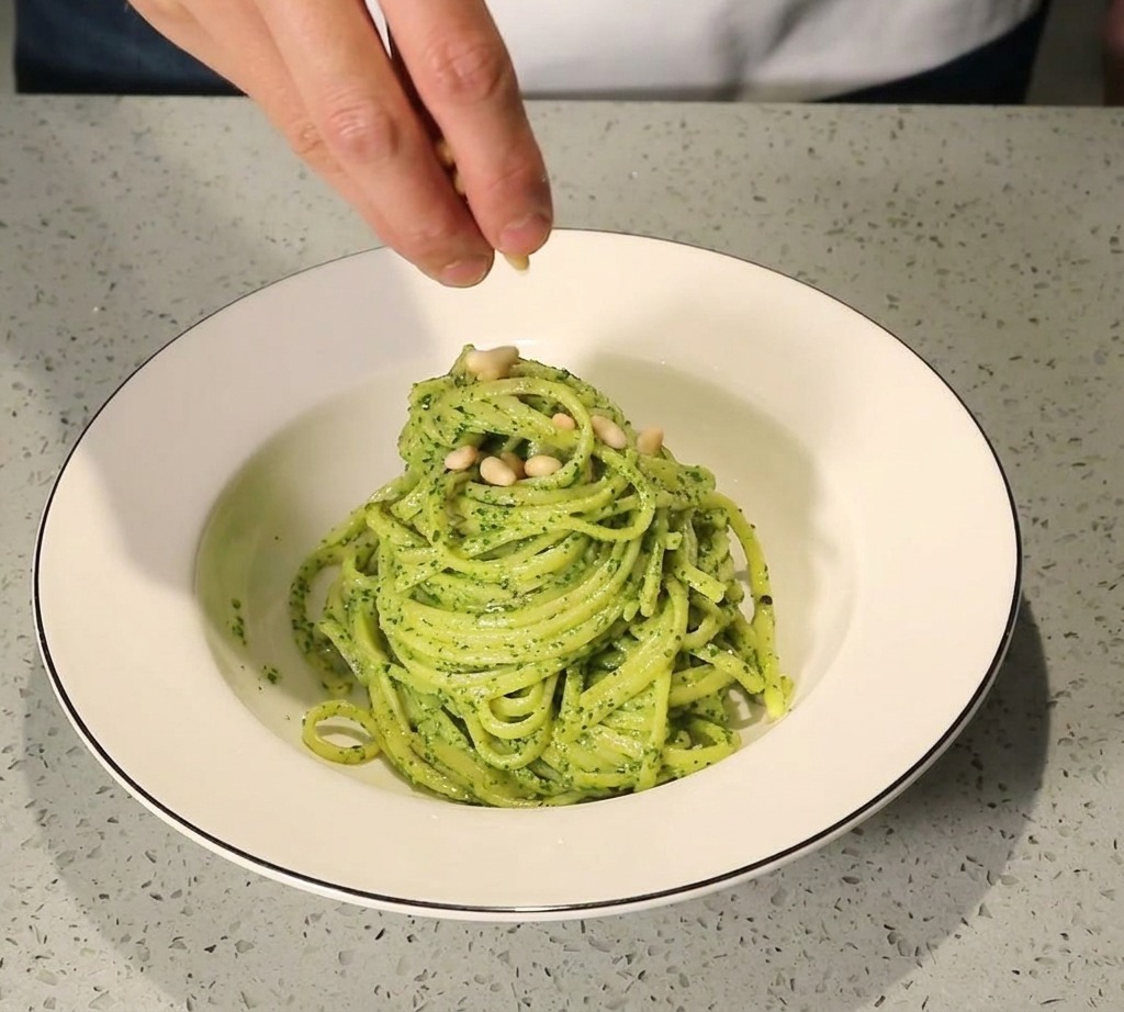 Una mano espolvoreando piñones tostados sobre una ración de pasta al pesto en un bol blanco.