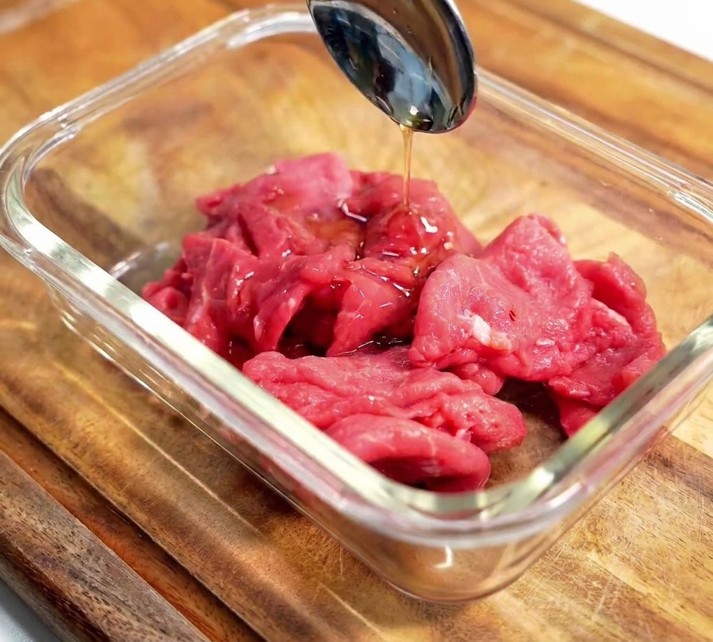 Una cuchara de metal vertiendo un condimento líquido oscuro sobre rodajas de carne cruda en un bol de cristal sobre una superficie de madera.