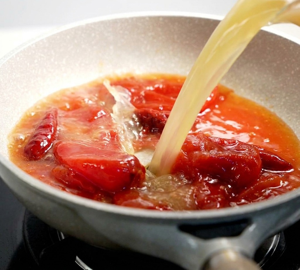 Un caldo de color claro vertiéndose en una sartén llena de una mezcla de salsa de tomate roja y burbujeante.