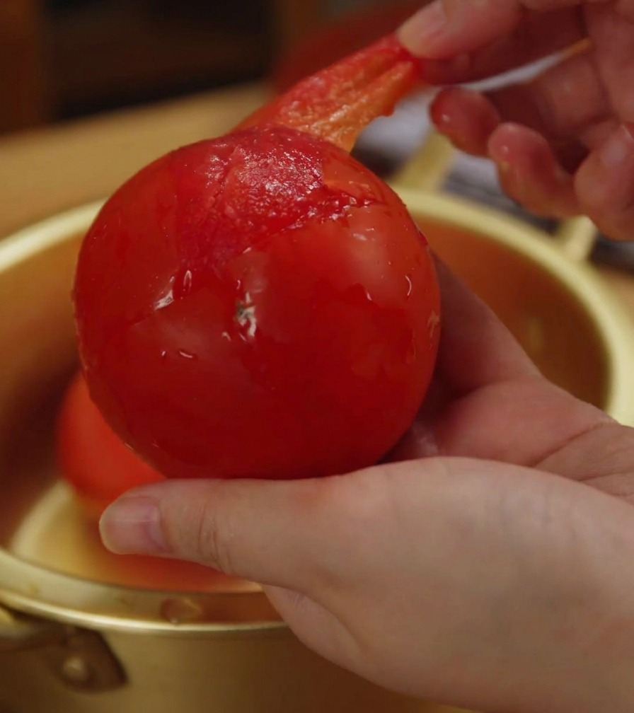 Primer plano de una mano retirando la piel fina y traslúcida de un tomate suavizado.