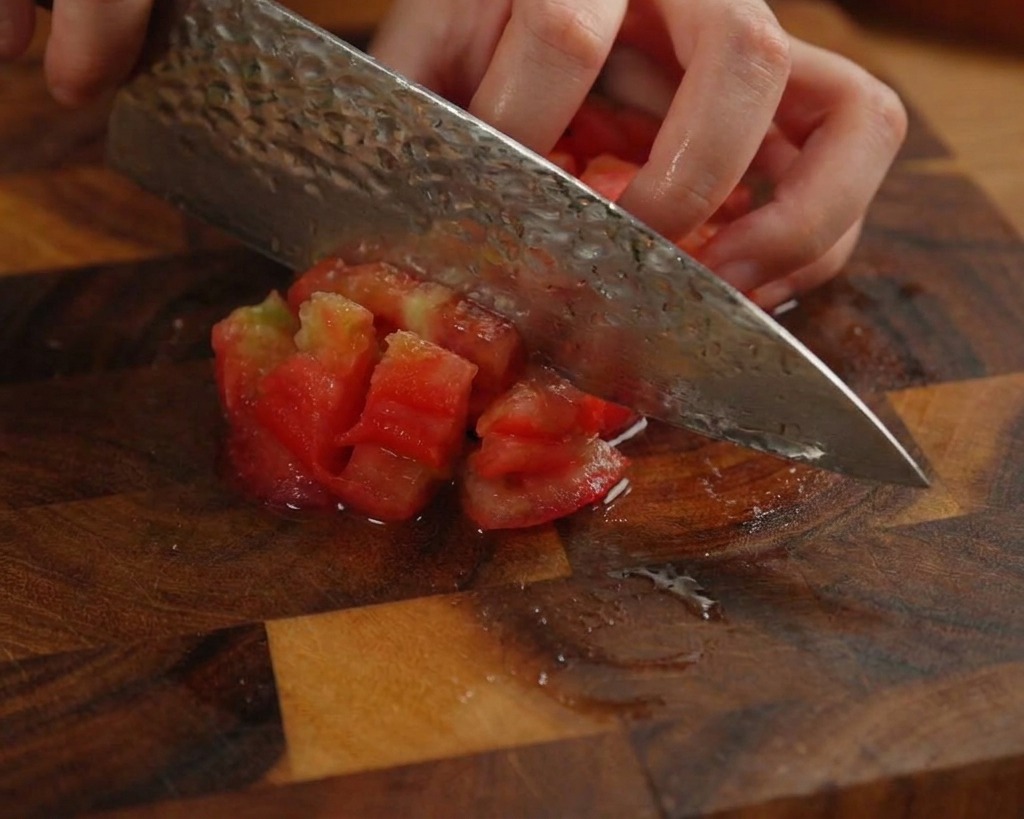 Una persona picando un tomate pelado en cubos pequeños sobre una tabla de madera oscura.