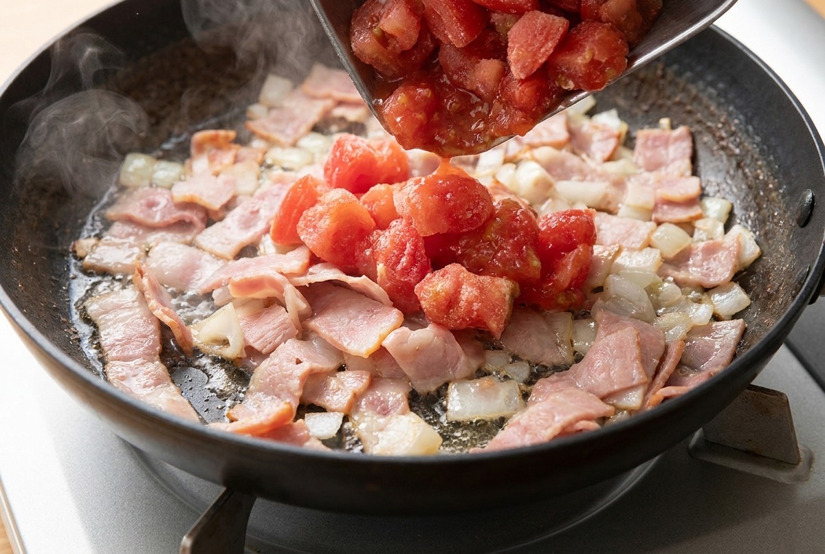 Tocino y cebolla picada cocinándose en una sartén mientras se añaden los tomates troceados.