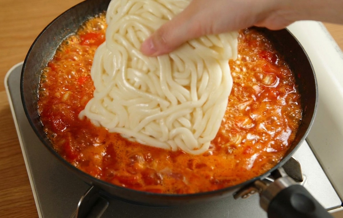 Una mano colocando un bloque cuadrado de fideos udon blancos en una sartén con salsa de tomate hirviendo.