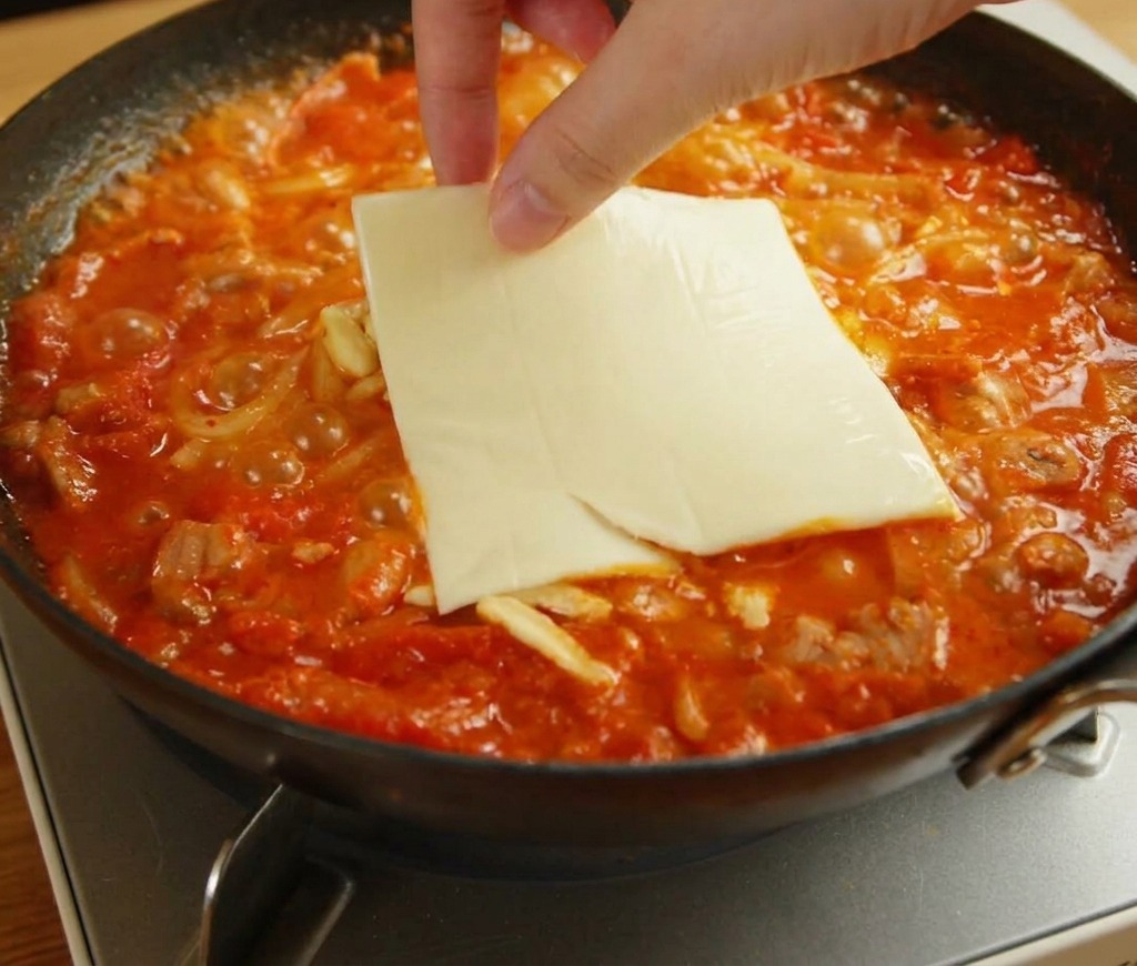 Mano colocando una rebanada cuadrada de queso amarillo sobre la mozzarella fundida y los fideos.