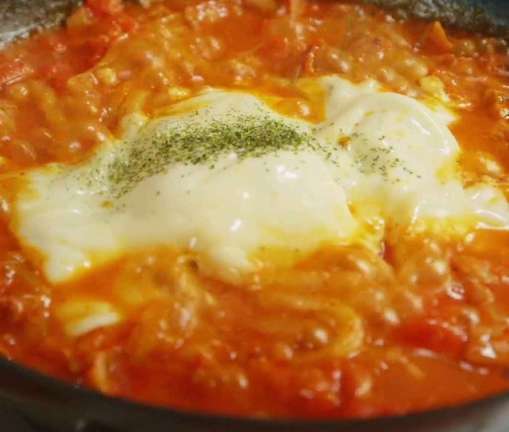 Hierbas verdes secas siendo espolvoreadas sobre una capa de queso blanco y amarillo fundido.