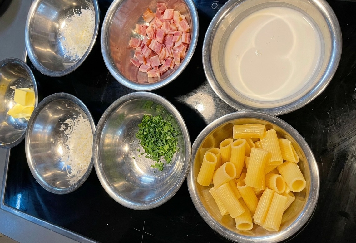 Varios cuencos de acero inoxidable con rigatoni, nata líquida, beicon picado, queso y perejil sobre una cocina negra.