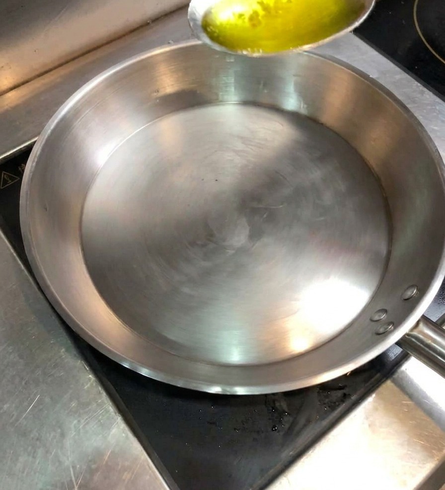 Cuchara de metal vertiendo aceite vegetal en una sartén de acero inoxidable caliente.