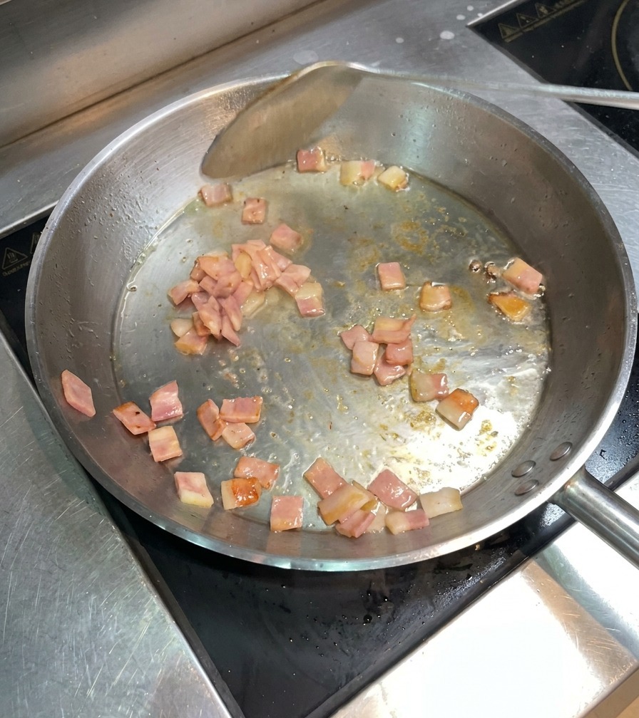 Trozos de beicon dorándose en la sartén mientras se remueven con una espátula metálica.