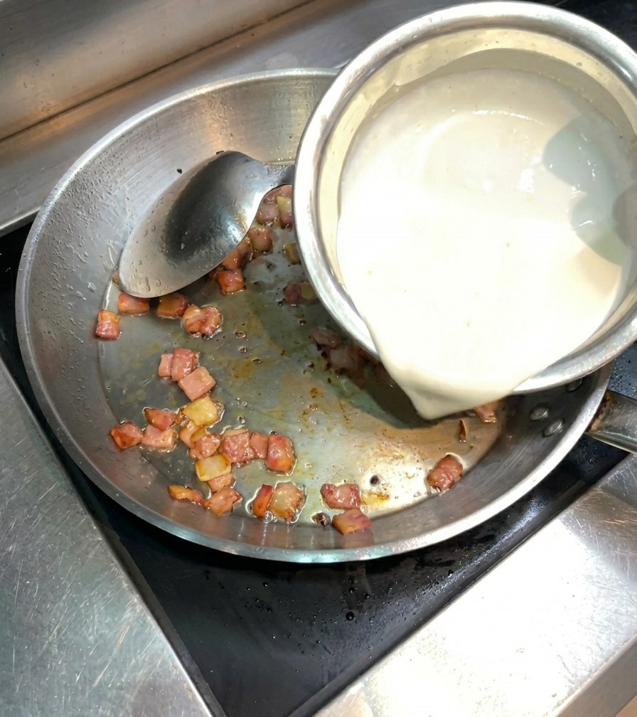 Nata espesa vertiéndose en la sartén de acero con el beicon salteado.