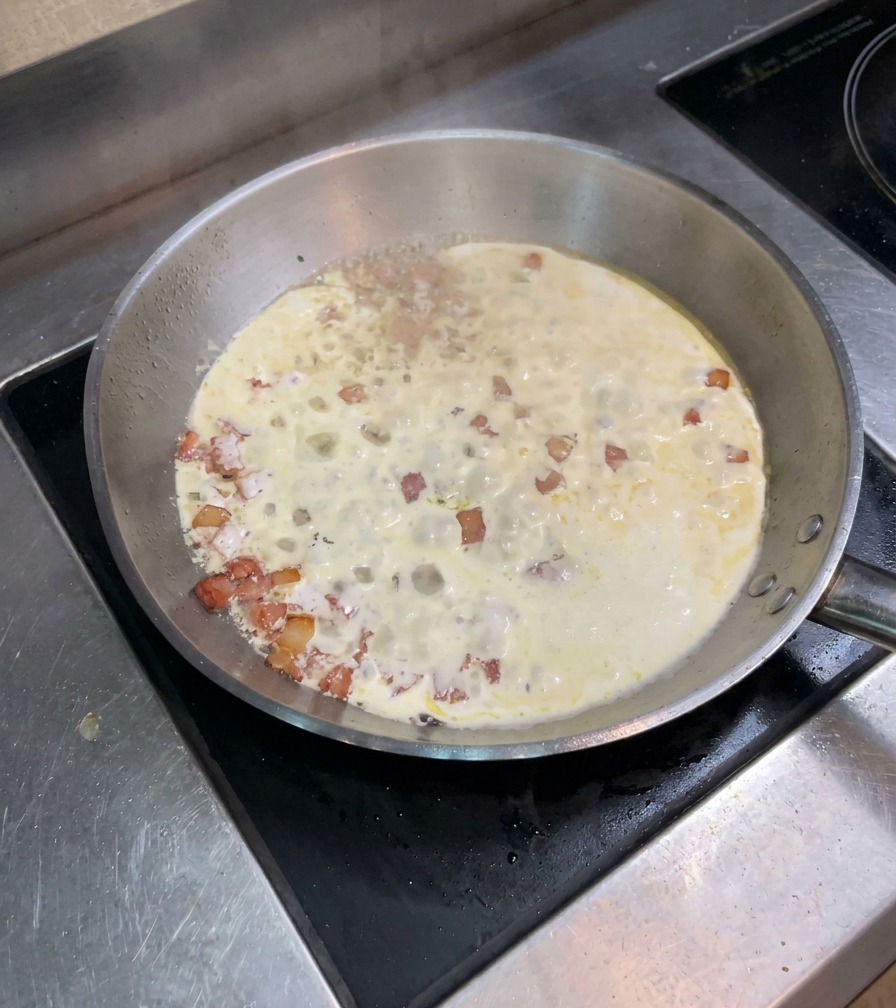 Mezcla de nata y beicon hirviendo suavemente en una sartén de acero inoxidable.