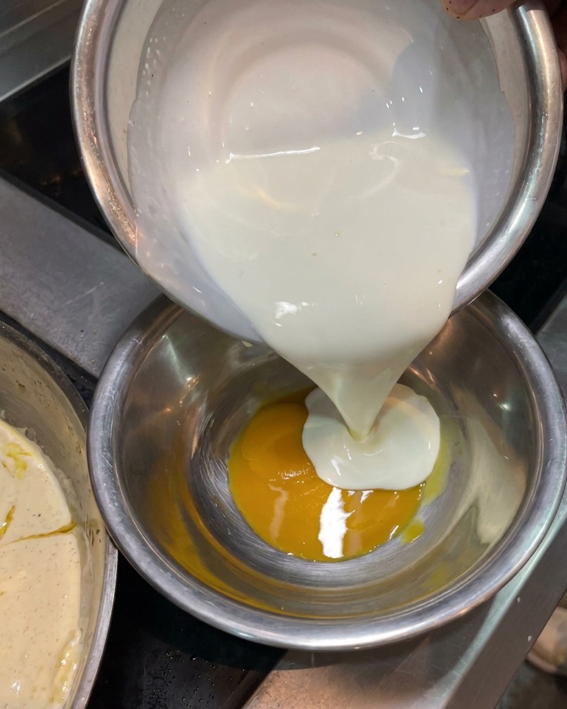 Nata vertiéndose desde un cuenco plateado a otro que contiene una yema de huevo amarilla.