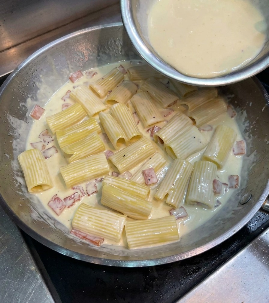 Mezcla amarilla de huevo y queso vertiéndose sobre los rigatoni en la sartén.
