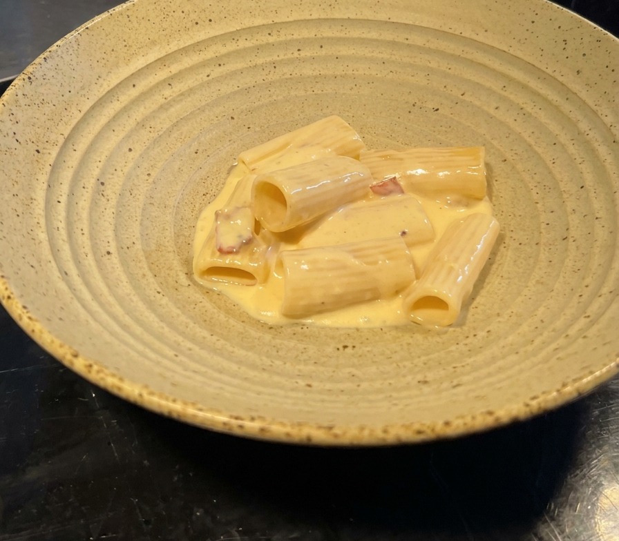 Ración de rigatoni con salsa de nata brillante y trozos de beicon en un cuenco cerámico.