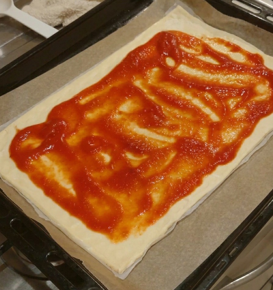 Una base de pizza rectangular con salsa de tomate roja extendida uniformemente, dejando un borde de masa limpio.