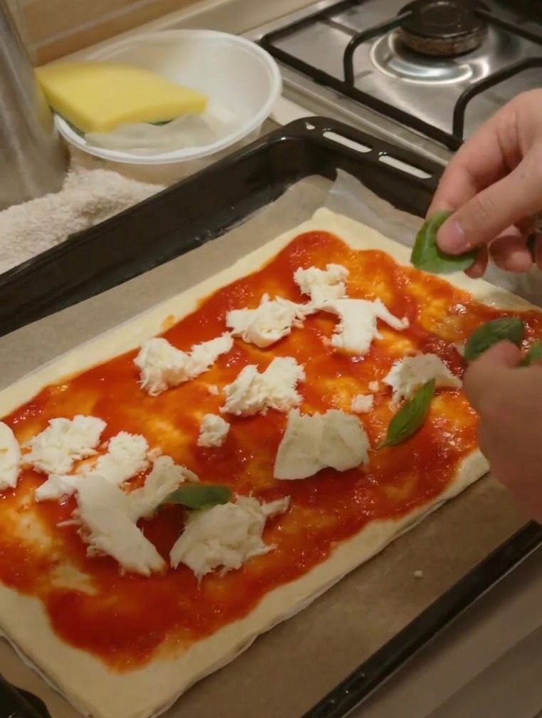 Manos colocando hojas de albahaca verde fresca sobre una pizza ya cubierta con salsa y trozos de queso.