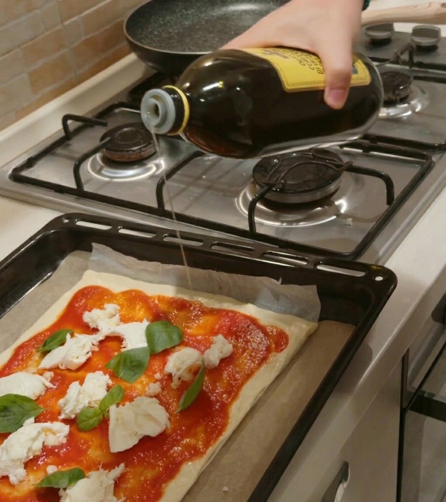 Una mano vertiendo un chorro de aceite de oliva desde una botella de cristal sobre una pizza cruda con salsa, mozzarella y albahaca.